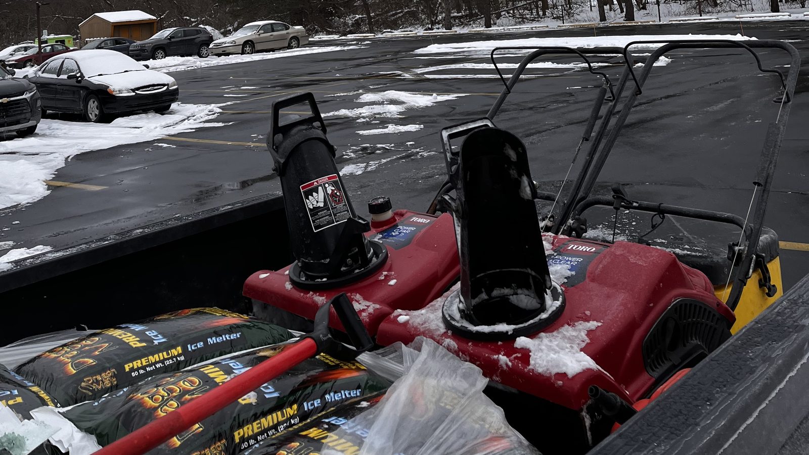 Bags of ice melter and a snow blower in the bed of a Freedom plow truck, with a snowy parking lot behind